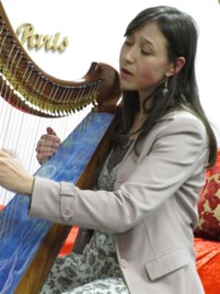 A woman with long hair plays a harp and sings.