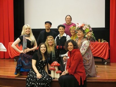 9 people dressed in fun casual clothes, 2 with floral headdresses, face the camera sitting around a statue of 3 red-capped mushrooms.